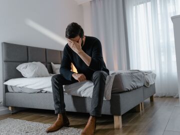 Un hombre reflexivo con un su&eacute;ter negro se sienta en una cama. 