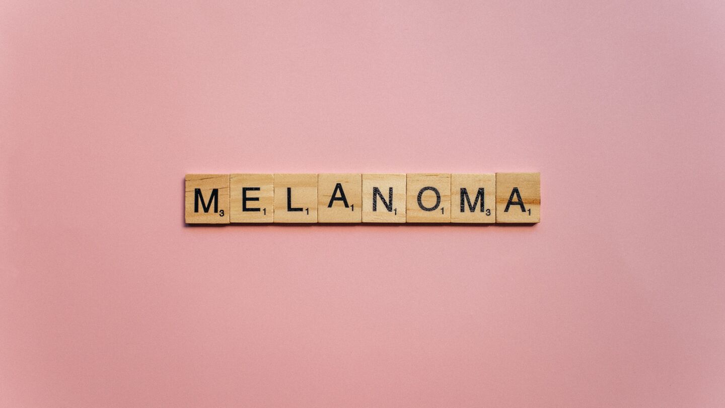 Letras de Scrabble de madera escribiendo 'Melanoma' sobre un fondo rosa suave, simbolizando la concienciación.