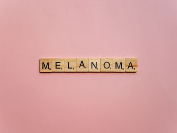 Letras de Scrabble de madera escribiendo 'Melanoma' sobre un fondo rosa suave, simbolizando la concienciaci&oacute;n.